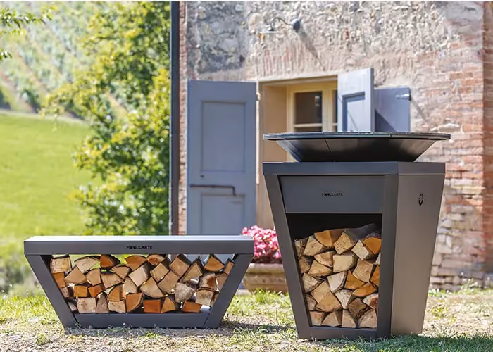 Plancha-Grill mit passender Holzunterstand-Bank in einem rustikalen Garten, vor einem Natursteinhaus.