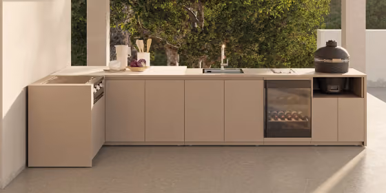 Lunga cucina da esterno color tortora sotto un portico, con lavello, cantinetta a vista e barbecue kamado nero, davanti a un albero
