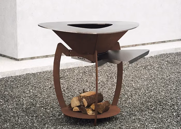 Moderner Außengrill mit Korpus in Corten-Optik, runder Kochplatte, seitlicher Stahlablage und Holzfach darunter, auf Kies stehend.