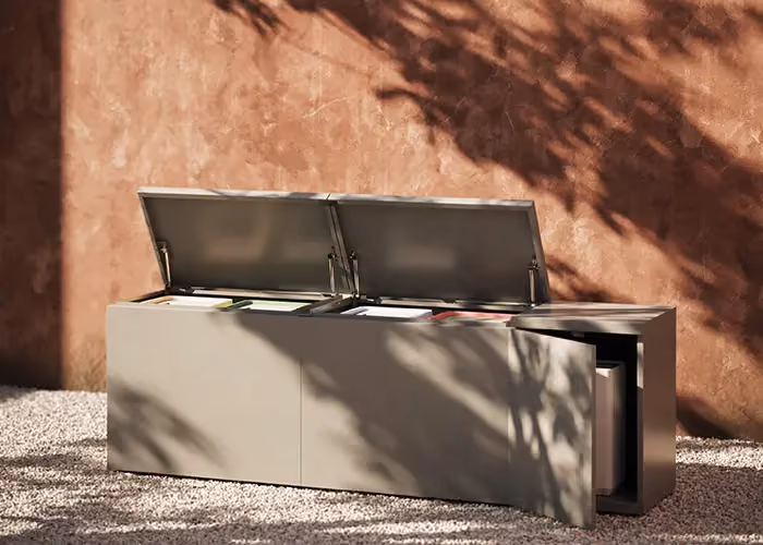 Modern outdoor storage unit with lift-up lids and an integrated side bay, standing on gravel in front of a terracotta-coloured wall.