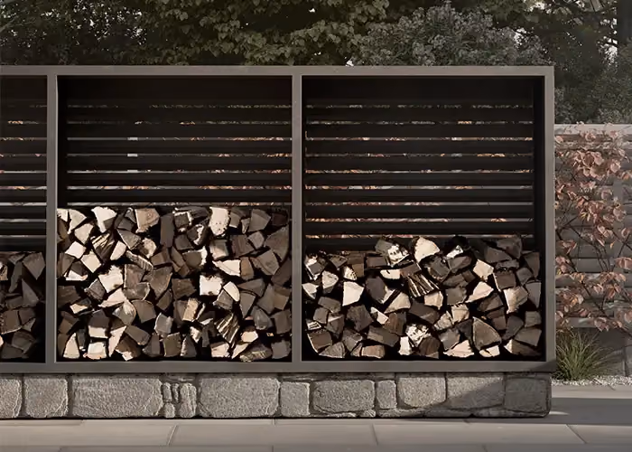 Modern outdoor log store in dark metal with horizontally slatted upper panels and lower bays filled with firewood, in front of a low stone wall.