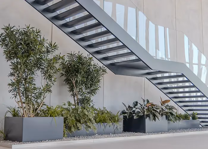 A run of grey rectangular planters with assorted green plants, set under a modern metal staircase.