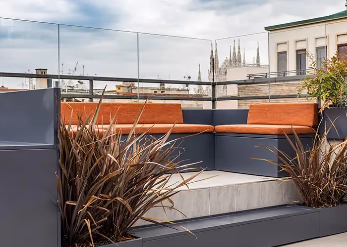 Modern lounge area on a city terrace with dark metal modular outdoor seating, orange cushions and integrated planters with ornamental planting.