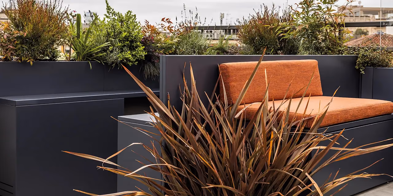 Outdoor sofa with a dark grey back and orange cushions, beside a planted planter