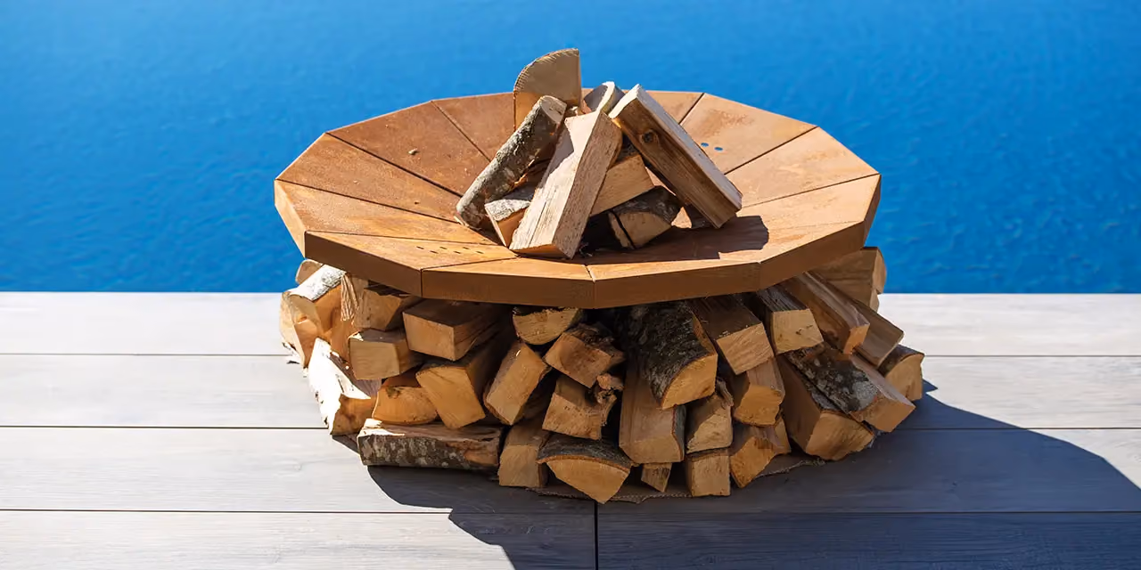 Circular wood and metal fire bowl on a jetty, loaded with logs, facing the sea