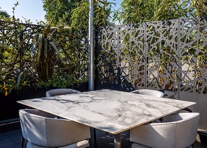 Marmor-Esstisch mit grauen Stühlen auf einem Patio, begrenzt von geometrischen Sichtschutzpaneelen aus Metall und Pflanzen.