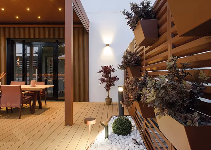 Patio moderne éclairé en soirée, avec sol en bois, pergola et bacs suspendus à un mur brise-vue.