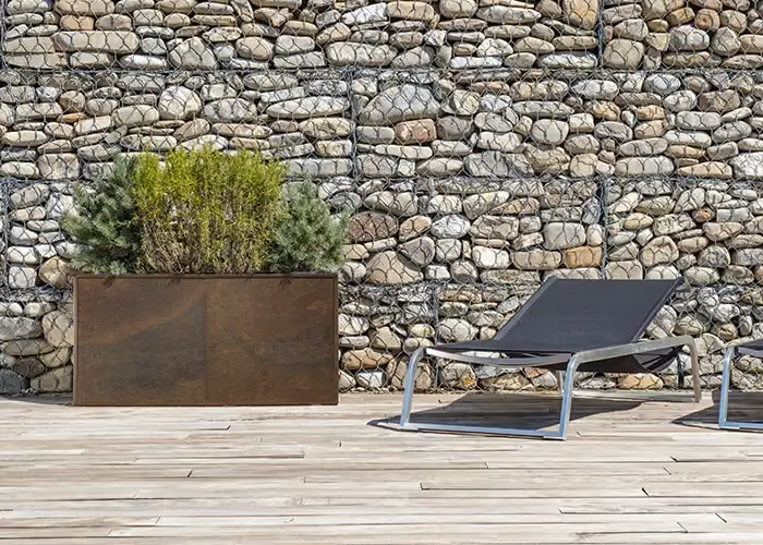 Rectangular corten-effect planter with ornamental shrubs, on a wooden terrace beside outdoor sun loungers.