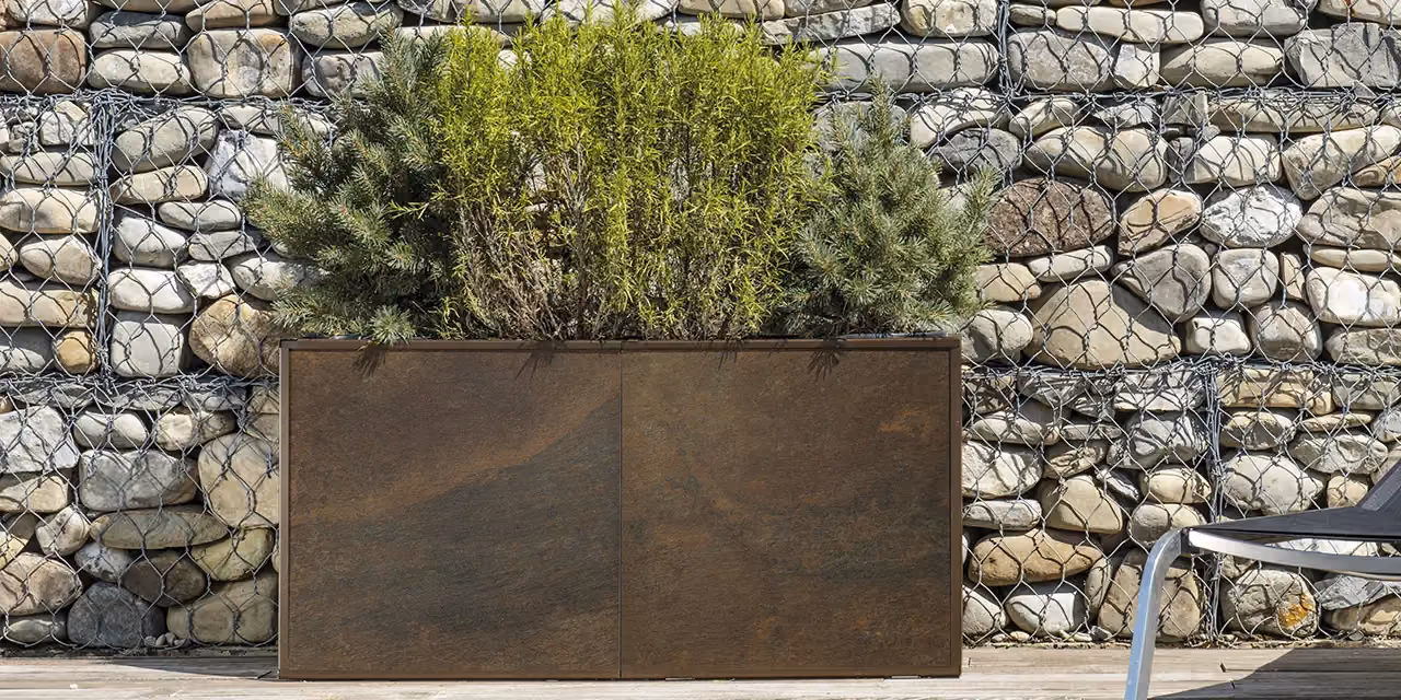 Rectangular corten steel planter against a stone wall, with shrubs