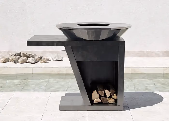 Modern outdoor barbecue in black metal with an integrated side surface, a lower log bay and a circular hearth, beside a swimming pool.