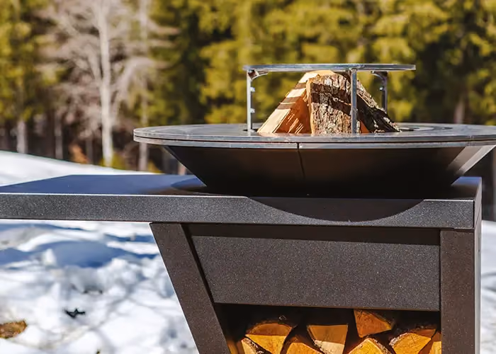 Modern outdoor barbecue with a circular hearth, a log rest and a lower log bay, set in a snowy mountain landscape.