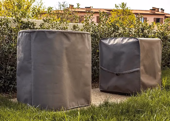 Grey protective covers for outdoor barbecues, set on a lawn beside a hedge.