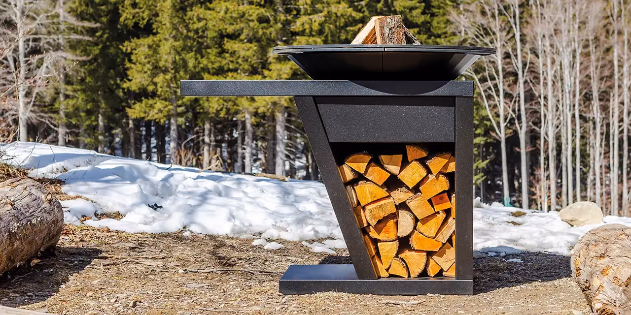 Tall black fire pit with an upper hearth and a lit log bay, in a snowy wood