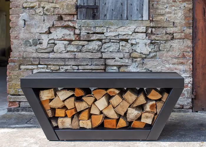Moderner Außen-Holzunterstand mit geometrischem Rahmen aus dunklem Metall und mit Brennholz gefülltem Mittelfach, vor einer rustikalen Wand aus Stein und Ziegel.