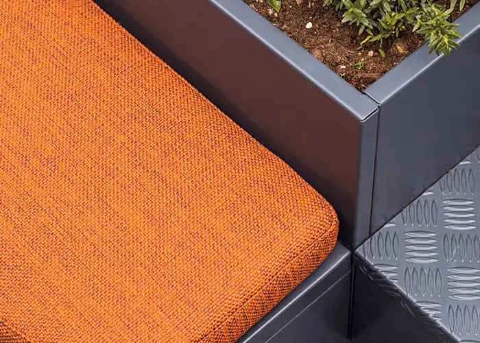 Detail einer modularen Außensitzbank mit orangefarbenem Kissen und integriertem Pflanzgefäß aus dunkelgrauem Metall mit Bepflanzung.