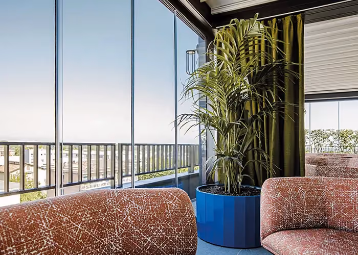 Ambiente lounge moderno con poltrone imbottite e grande fioriera blu contenente una palma ornamentale vicino a vetrate panoramiche.