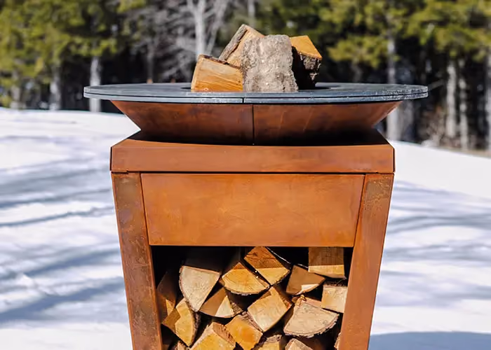 Runder Grill aus rostfarbenem Cortenstahl mit Brennholz, in einer verschneiten Landschaft.