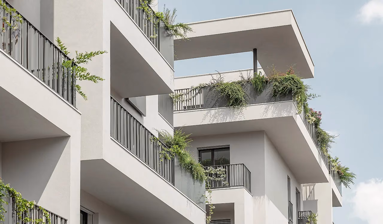 Upward view of modern corner balconies and terraces on a white building, with grey perimeter planters and green plants trailing from them.