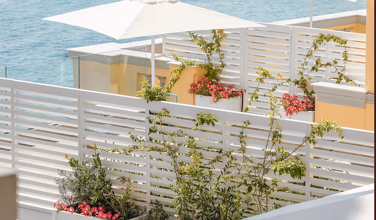 Seafront terrace with white louvred wooden screening panels, white rectangular planters with pink flowers and a parasol.