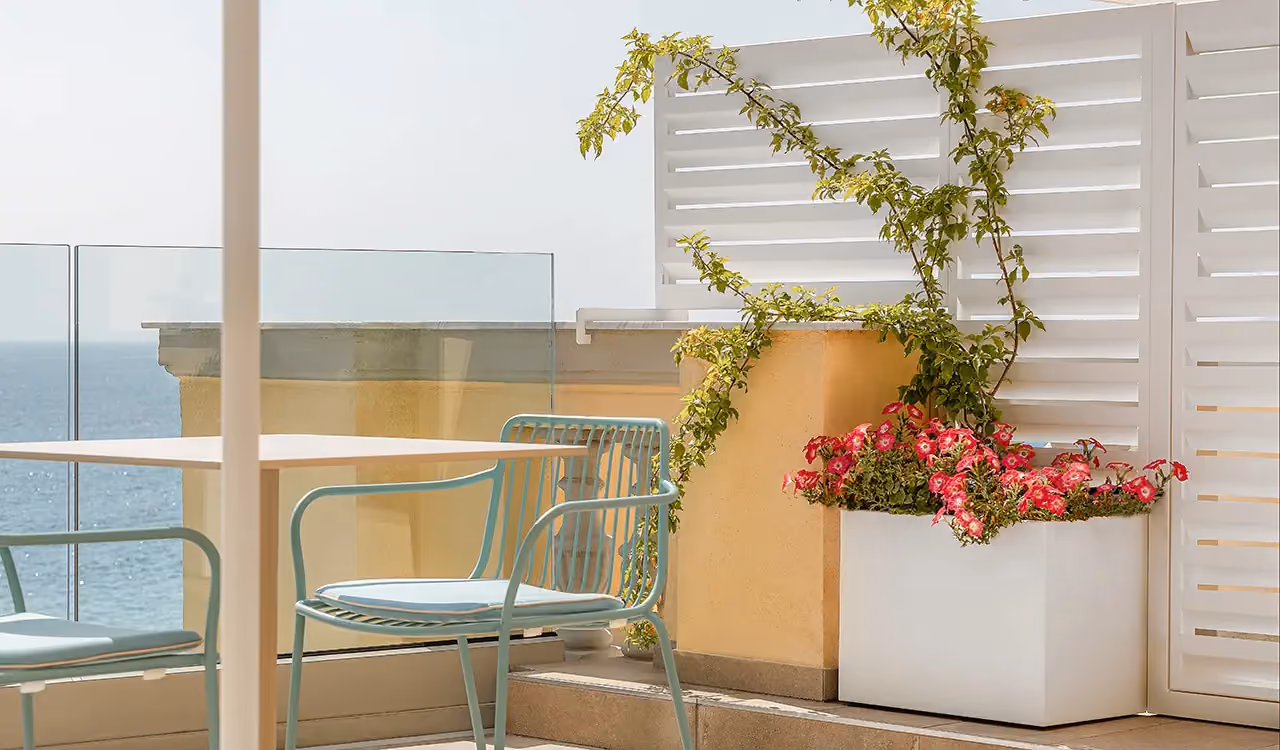 Detail of a sea-view terrace with a light blue metal chair, a side table, a white planter with pink bougainvillea and a white windbreak panel behind.