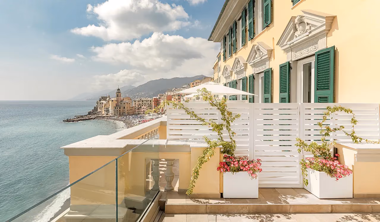 Wide panoramic terrace of a yellow historic building overlooking a bay and a coastal village, bounded by white screening panels with planters.